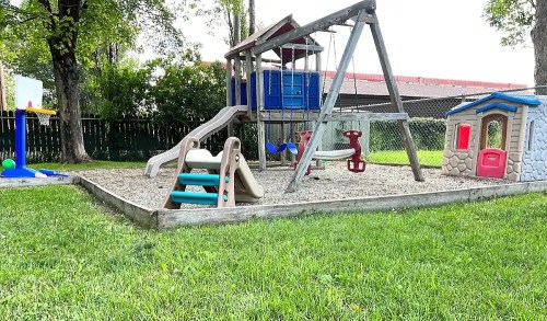 A playground with a slide, swings, and a playhouse, surrounded by grass and trees.