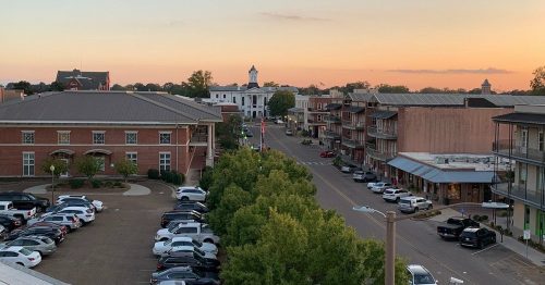 A sunset view of a small town street lined with buildings and parked cars, with trees in the foreground.