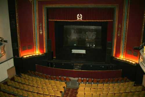 A view of an empty theater stage with red curtains and rows of yellow seats in the foreground.