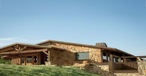 A modern stone and wood house with a sloped roof, set against a clear blue sky and green grass.