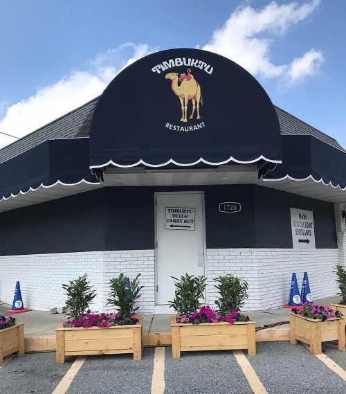 Exterior of Timbuktu Restaurant featuring a camel logo, blue awning, and flower planters at the entrance.