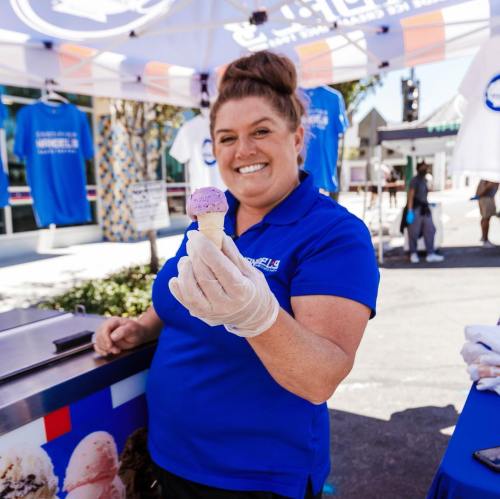 A smiling woman in a blue shirt holds a cone of purple ice cream at a festive outdoor event.