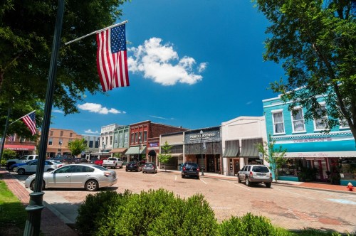 A charming street scene with colorful storefronts, American flags, and clear blue skies. Cars are parked along the road.