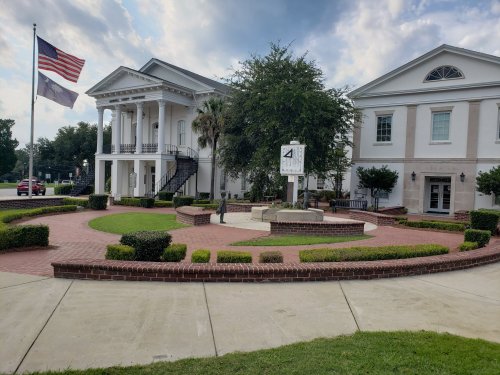 A historic building with columns, surrounded by landscaped gardens and a flagpole, featuring a clock in the center.
