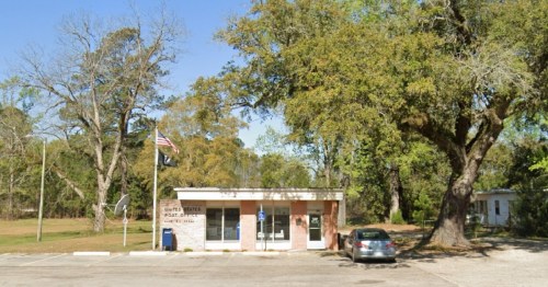 A small post office building with an American flag, surrounded by trees and a parking area.