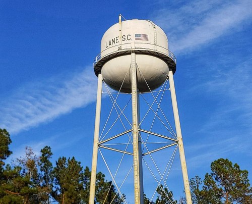 A white water tower labeled "LANE S.C." stands tall against a blue sky with scattered clouds and trees below.