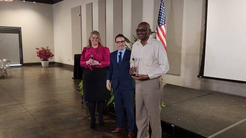 Three individuals stand together on stage, holding awards, with an American flag in the background.