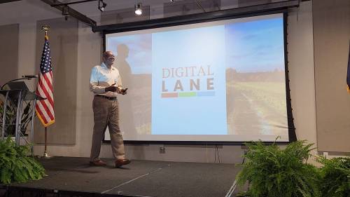 A speaker stands on stage in front of a screen displaying "DIGITAL LANE," with greenery in the foreground.
