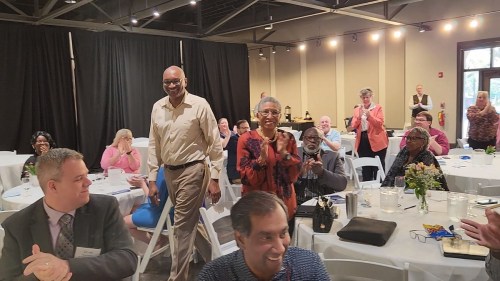 A diverse group of people applauding as two individuals walk towards the front of a gathering in a well-lit venue.