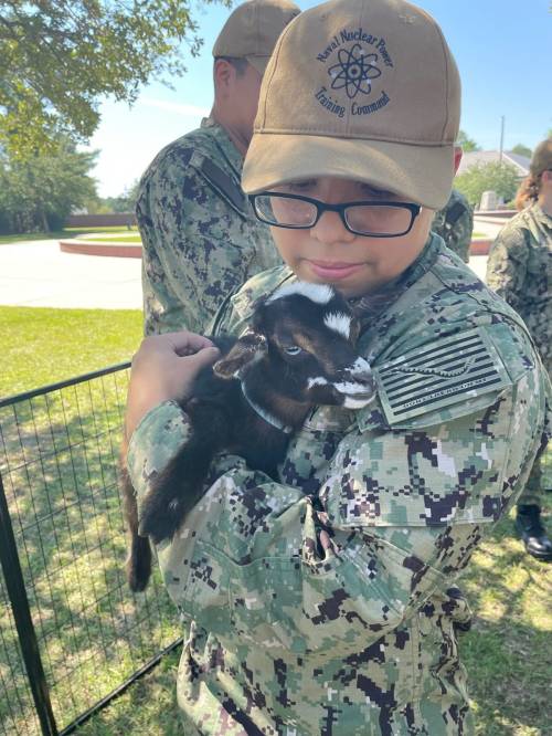 A child in military camouflage holds a small goat, wearing a cap, surrounded by others in similar attire outdoors.
