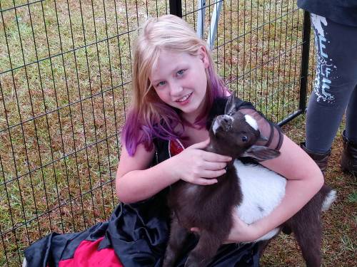 A girl with purple-tipped hair sits on the grass, holding a small goat in her arms, smiling at the camera.