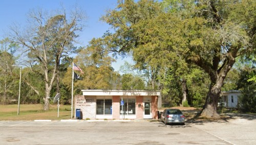 A small brick post office building with an American flag, surrounded by trees and a parking area.