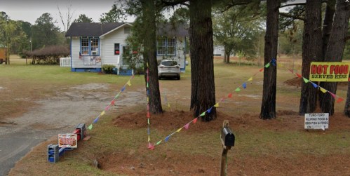 A roadside eatery with a sign for hot food, colorful flags, and a parked car in a rural setting.