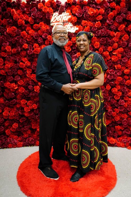 A couple stands together in front of a wall of red roses, both smiling and dressed in vibrant, traditional attire.