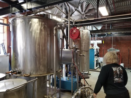 A person works in a brewery with large stainless steel tanks and brewing equipment in a spacious industrial setting.