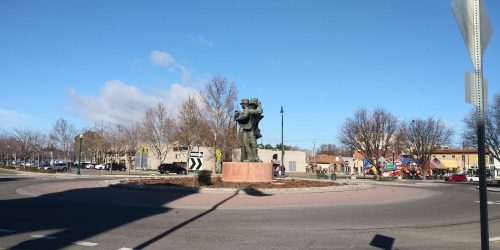 A large statue in a roundabout, surrounded by trees and buildings under a clear blue sky.