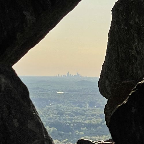 A view of a distant city skyline framed by rocky formations, with greenery in the foreground and a hazy sky.