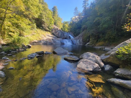 A serene waterfall cascades into a clear pool, surrounded by lush greenery and rocky terrain under a blue sky.