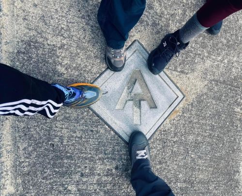 Three pairs of shoes standing around a metal marker with the letter "A" on a concrete surface.