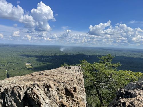 A rocky cliff overlooks a lush green valley under a blue sky filled with fluffy white clouds.