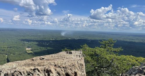 A panoramic view from a rocky outcrop, showcasing lush green forests and a blue sky with fluffy clouds.