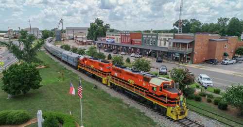 A colorful train passes by a small town with shops, trees, and flags under a cloudy sky.