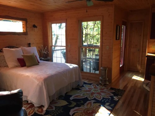 Cozy bedroom with a white bed, colorful rug, wooden walls, and large windows overlooking a deck and trees.