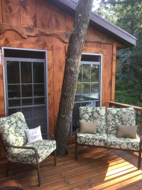 A cozy outdoor seating area with floral chairs beside a wooden cabin, featuring a tree growing through the deck.