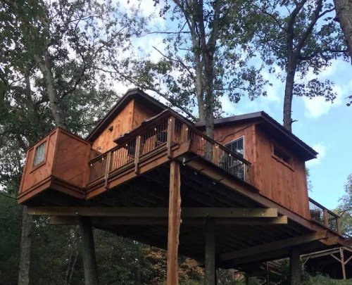 A wooden treehouse elevated on stilts, surrounded by trees, with a person standing beside it on the ground.