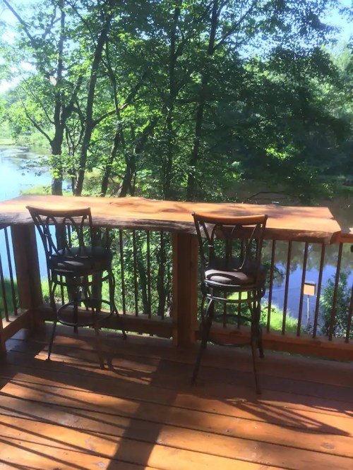 A wooden deck with two bar stools overlooking a serene river and lush greenery.