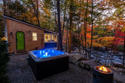 A cozy cabin by a river, featuring a hot tub and a fire pit, surrounded by vibrant autumn foliage.