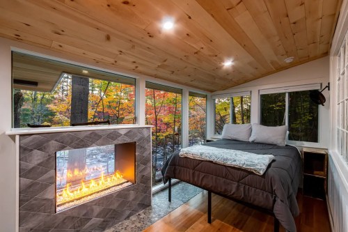 Cozy bedroom with a modern fireplace and large windows showcasing vibrant autumn foliage outside.