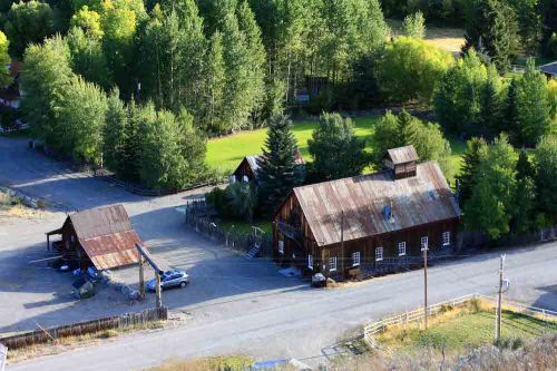 Aerial view of a rustic wooden building and shed surrounded by trees and a grassy area, near a gravel road.