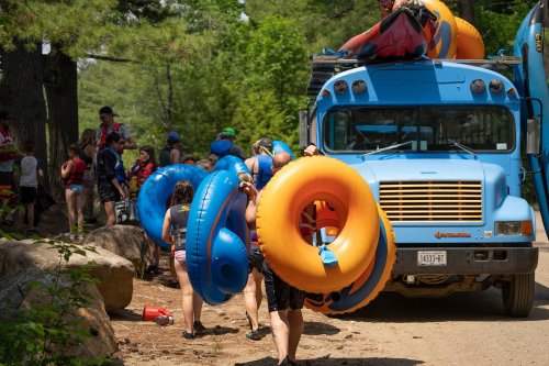 People carrying colorful inner tubes near a blue truck, surrounded by trees and a group preparing for water activities.