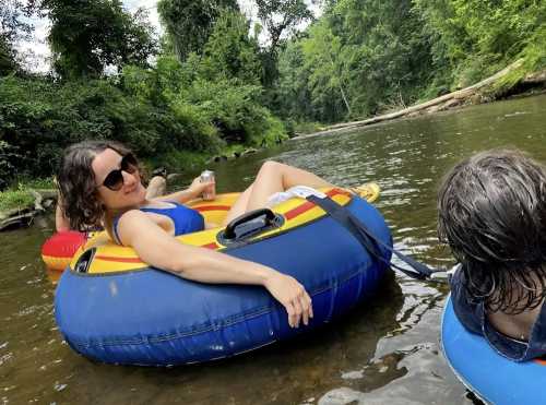 A woman in sunglasses relaxes on a blue inner tube in a river, surrounded by lush greenery. Two others are nearby.