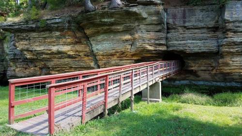 A wooden bridge leads to a cave entrance in a rocky cliff, surrounded by green grass and trees.