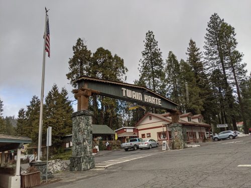 Entrance sign for Twain Harte, surrounded by tall trees and buildings, with an American flag in the background.