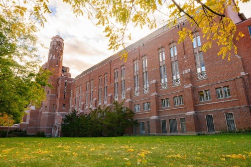 Historic brick building with tall tower, surrounded by green grass and autumn leaves under a cloudy sky.