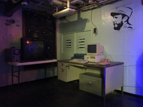 A dimly lit room featuring an old TV, a vintage computer, a desk, and a wall mural of a man’s profile.