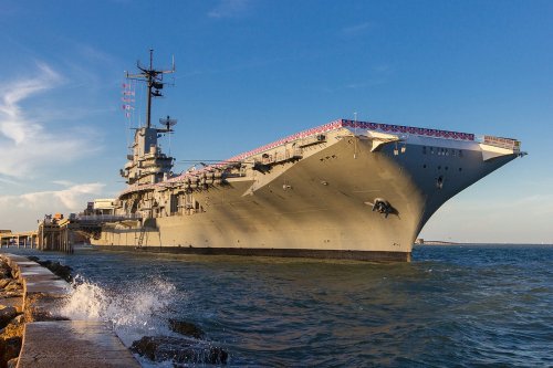 A large aircraft carrier docked at a pier, with waves splashing against the side and a clear blue sky above.