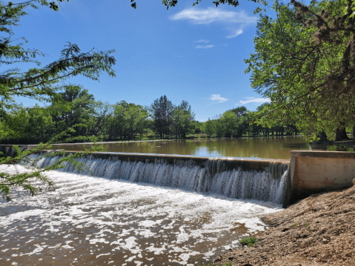 A serene landscape featuring a waterfall cascading over a dam, surrounded by lush greenery and a clear blue sky.