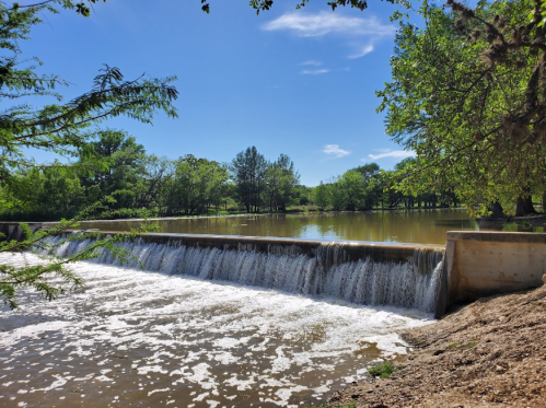 A serene landscape featuring a waterfall cascading over a dam, surrounded by lush greenery and a clear blue sky.