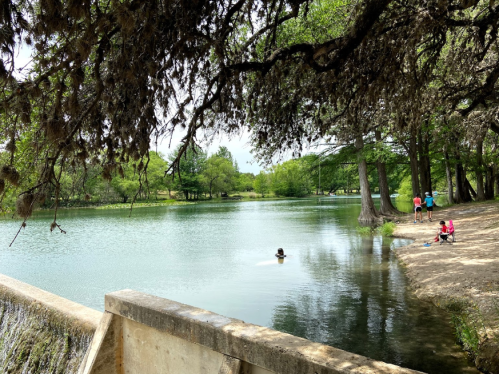 A serene park scene with a calm lake, trees, and people enjoying the outdoors by the water's edge.