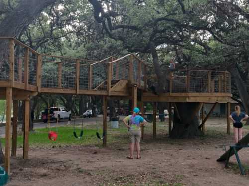 A wooden treehouse with a slide and swings, surrounded by trees, with two people observing nearby.