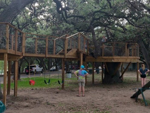 A wooden treehouse with a slide and swings, surrounded by trees, with two people observing nearby.