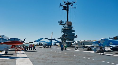 A naval aircraft carrier deck with various planes and personnel under a clear blue sky.