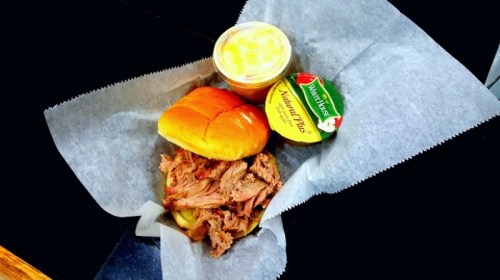 A basket with pulled pork, a soft bun, a cup of fruit, and a small container of sauce, all on parchment paper.