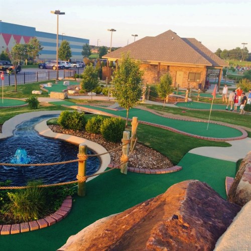 A scenic mini-golf course with a pond, greenery, and a clubhouse in the background during sunset.