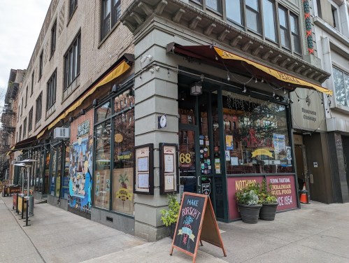 A street view of a restaurant with large windows, outdoor seating, and a colorful sign on the sidewalk.
