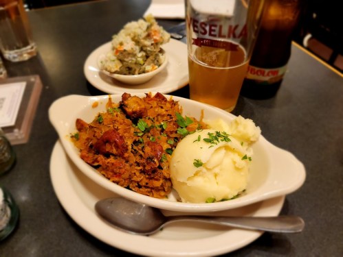 A plate of hearty meat dish with mashed potatoes, a side of coleslaw, and a glass of beer on a table.